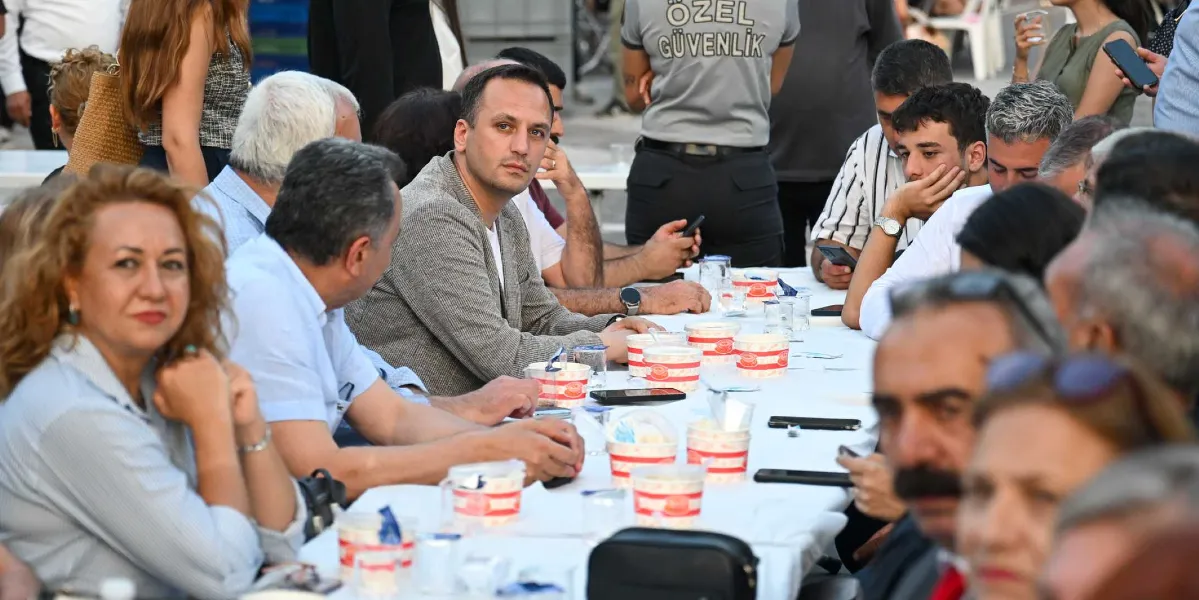 Açık havada uzun bir masanın etrafında toplanmış, önlerinde aşure kaseleri bulunan kalabalık bir grup insan. Masanın orta kısmında gri ceketli bir adam doğrudan kameraya bakarken, diğer katılımcılar sohbet ediyor veya telefonlarıyla ilgileniyor. Arka planda üzerinde 
