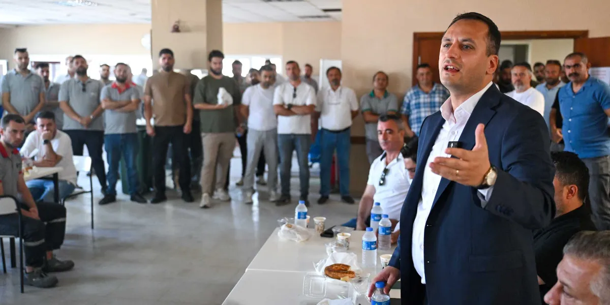 Koyu renk ceket ve beyaz gömlek giymiş bir erkek, kapalı bir alanda ayakta durarak konuşma yapıyor ve el hareketleriyle bir şeyler anlatıyor. Önünde üzerinde su şişeleri ve yiyecek kapları bulunan beyaz bir masa yer alıyor. Arka planda ise onu dinleyen, çoğunluğu erkeklerden oluşan kalabalık bir grup ayakta duruyor.