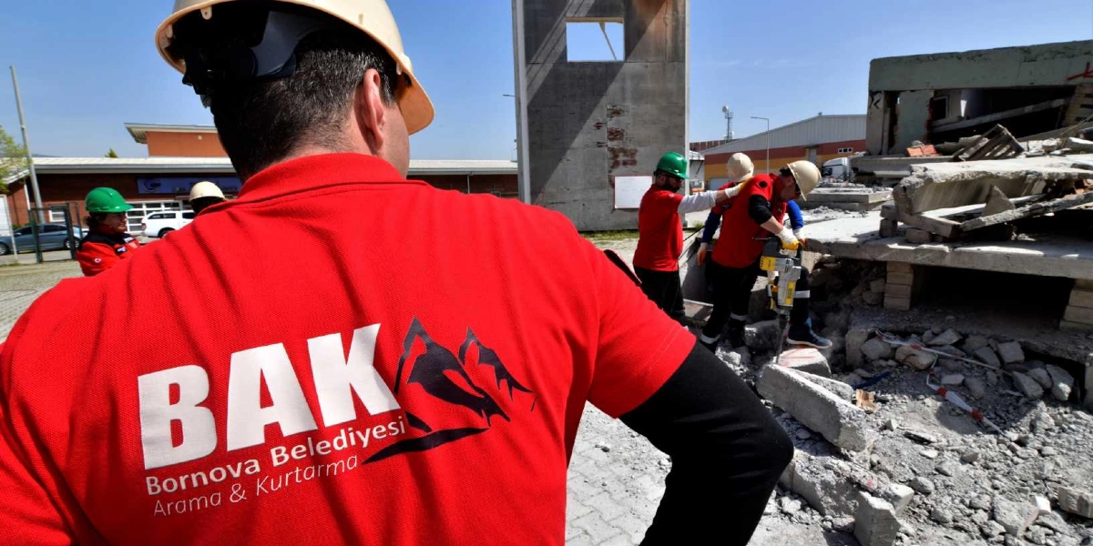 Üzerinde Bornova Belediyesi Arama ve Kurtarma (BAK) yazılı kırmızı üniformalı ve baretli ekip üyeleri, enkaz benzeri bir alanda arama kurtarma tatbikatı veya çalışması gerçekleştiriyor. Ekip üyelerinden biri elindeki kırıcı delici aletle beton parçaları üzerinde işlem yaparken, diğerleri onu gözlemliyor.