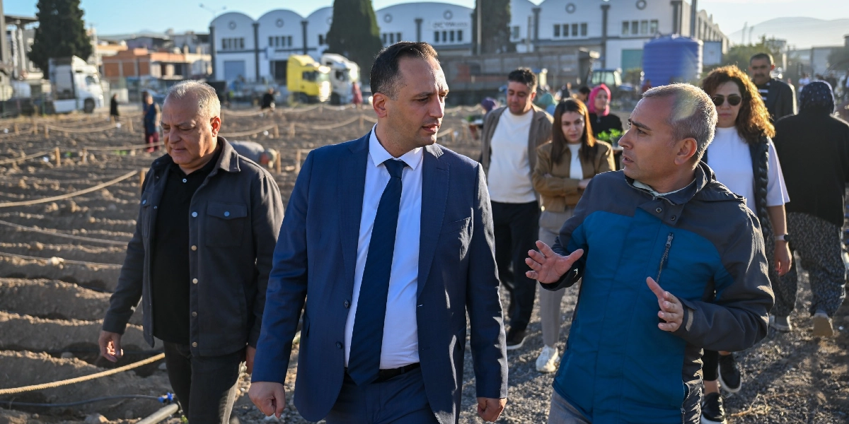 Açık havada, toprakla çevrili bir alanda yürüyen bir grup insan; ön planda lacivert takım elbiseli bir adam, yanındaki spor ceketli bir kişiyle konuşarak ilerliyor. Arka planda ise tarım alanı olduğu anlaşılan, iplerle bölümlere ayrılmış arazide çalışan diğer insanlar ve sanayi tipi yapılar görülüyor.