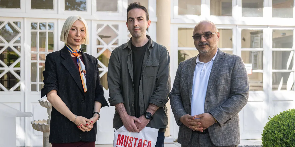 Tarihi beyaz bir binanın önünde yan yana duran üç kişi; solda sarı kısa saçlı, siyah ceketli ve boynunda renkli bir fular olan kadın, ortada haki renkli gömlek giyen ve elinde üzerinde kırmızı harflerle 