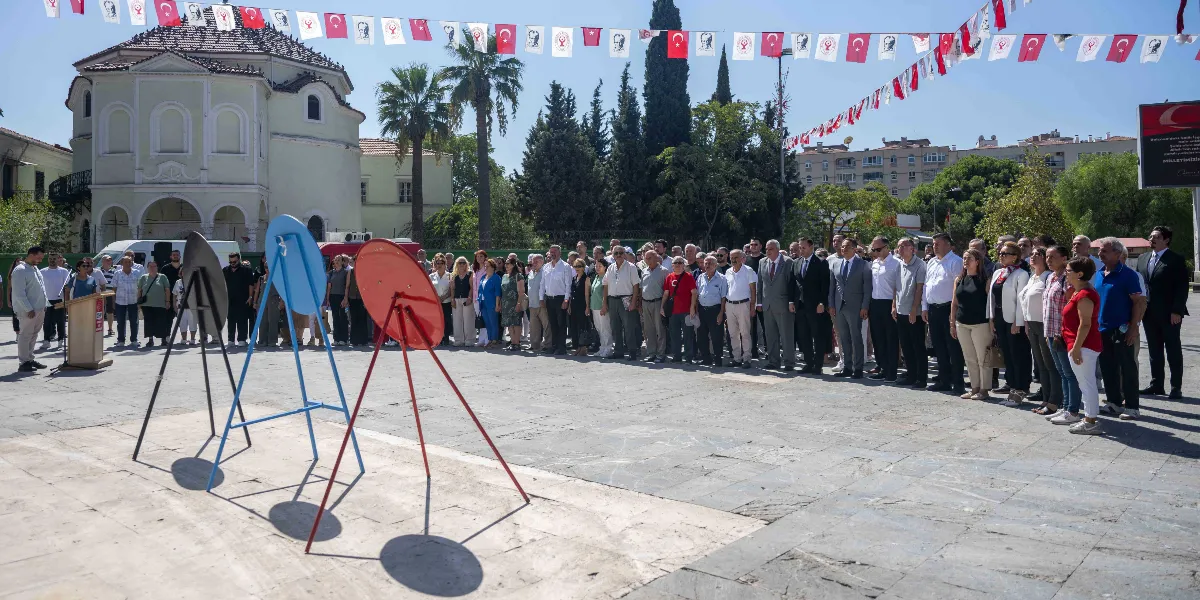 Bornova'da düzenlenen 9 Eylül töreninde, meydanda toplanmış kalabalık bir grup insan saygı duruşunda bulunuyor. Meydanın ön kısmında üç ayaklı sehpalara yerleştirilmiş boş çelenkler yer alırken, arka planda tarihi bir bina, ağaçlar ve Türk bayraklarıyla süslenmiş bir alan görülüyor.