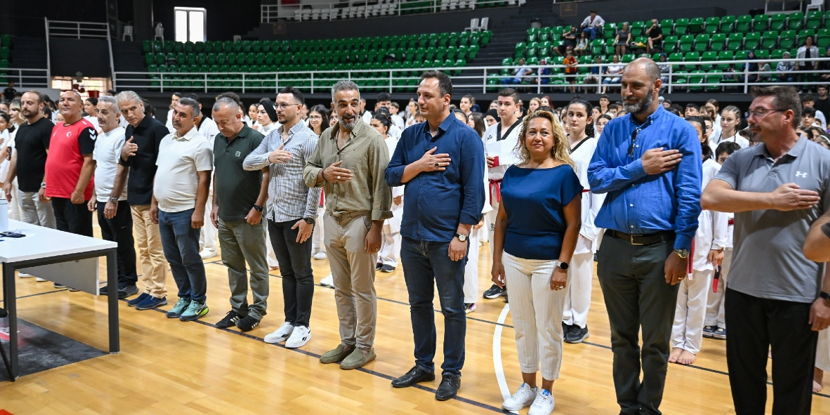 Kapalı bir spor salonunda, ellerini kalplerinin üzerine koyarak saygı duruşunda bulunan bir grup yetişkin ve arka planda beyaz tekvando kıyafetleri giymiş çok sayıda sporcu adayı yer alıyor. Ön sırada yan yana dizilmiş, günlük kıyafetler içindeki kadın ve erkeklerden oluşan protokol üyeleri, ciddiyetle karşıya bakıyor. Arka planda ise yeşil koltuklu tribünlerde oturan izleyiciler görülüyor.