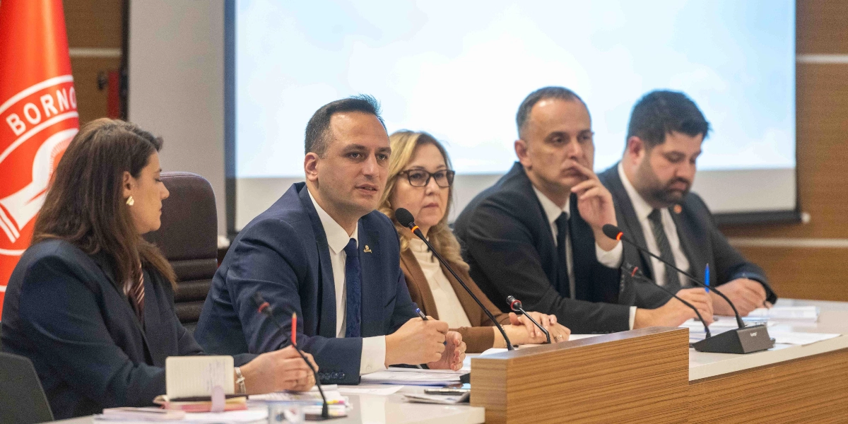 Toplantı masasında yan yana oturan, önlerinde mikrofonlar bulunan Bornova Belediyesi yetkilileri bir sunum veya toplantı esnasında konuşma yapıyor. Arka planda Bornova Belediyesi logosunun yer aldığı kırmızı bir bayrak ve projeksiyon perdesi bulunuyor.