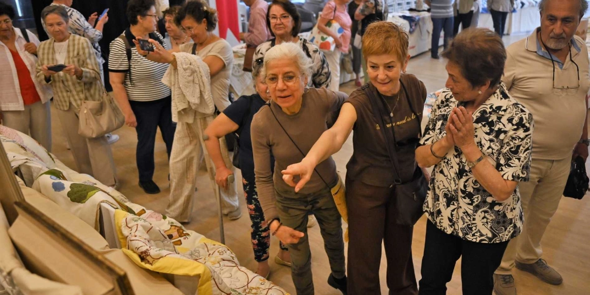 Atatürkçü Düşünce Derneği üyelerinin katıldığı bir sergide, el emeği ürünlerin sergilendiği stantları inceleyen bir grup insan. Ön planda, kahverengi tişörtlü bir kadın el işi ürünleri işaret ederek yanındaki arkadaşına gösteriyor, yanında ise ellerini birleştirmiş heyecanla bakan başka bir kadın bulunuyor. Arka planda ise ürünleri inceleyen, fotoğraflarını çeken ve sohbet eden katılımcılar yer alıyor.
