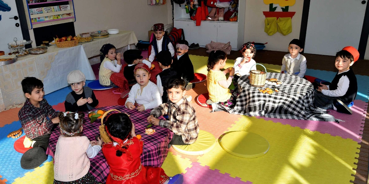 Yerli Malı Haftası kutlaması kapsamında anaokulu sınıfında geleneksel kıyafetler giymiş çocuklar, yer minderleri üzerinde kurulmuş yer sofralarının etrafında oturuyor. Masaların üzerinde meyveler ve yiyecekler bulunurken, çocuklar neşeyle bir arada vakit geçiriyor. Arka planda ise üzerinde çeşitli yiyeceklerin sergilendiği uzun bir masa ve çocukların eşyalarının bulunduğu dolaplar yer alıyor.