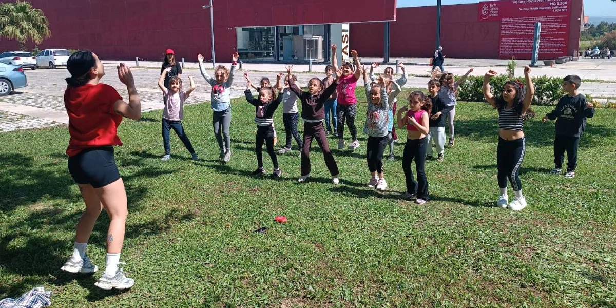 Yeşilova Höyüğü Ziyaretçi Merkezi'nin önündeki çim alanda, kırmızı tişörtlü bir eğitmen eşliğinde spor yapan bir grup çocuk. Çocuklar eğitmeni takip ederek kollarını havaya kaldırmış veya çeşitli hareketler yapıyor. Arka planda müze binası ve otopark alanı görülüyor.