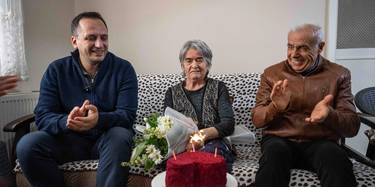 Ev ortamında, desenli bir koltukta yan yana oturan üç kişi; ortada oturan yaşlı bir kadın, kucağında beyaz çiçeklerden oluşan bir buket tutuyor. Kadının önündeki masada üzerinde yanan mumlar bulunan kırmızı bir pasta duruyor. Kadının sağında lacivert kazaklı, solunda ise kahverengi deri ceketli bir adam oturuyor ve her ikisi de gülümseyerek alkışlıyor.