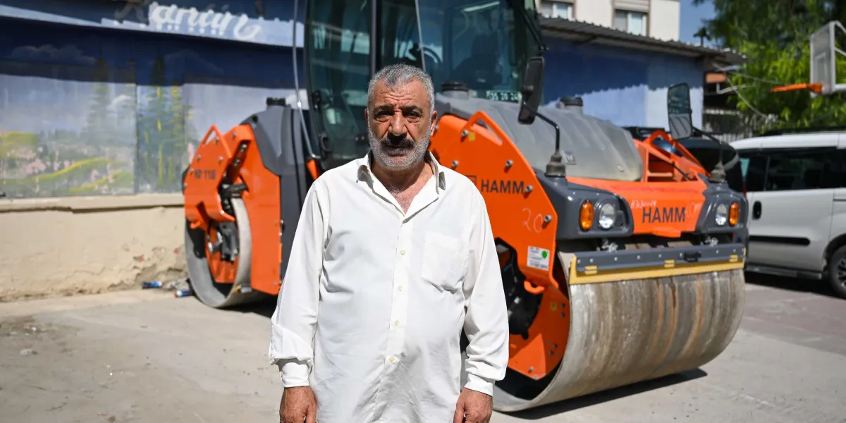 Beyaz gömlekli, bıyıklı orta yaşlı bir adam, arkasında duran turuncu renkli Hamm marka bir silindir iş makinesinin önünde duruyor. İş makinesinin üzerinde, saldırı sonucu oluştuğu anlaşılan belirgin bir hasar izi görülüyor.