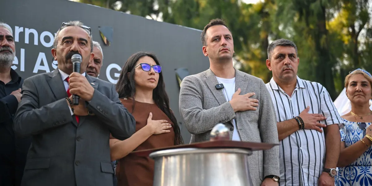 Açık havada düzenlenen bir etkinlikte, ellerini kalplerinin üzerine koyarak saygı duruşunda bulunan bir grup insan yer alıyor. En önde, elinde mikrofon tutan gri takım elbiseli bir adam konuşma yaparken, yanında kahverengi bluzlu bir kadın, gri ceketli bir erkek, çizgili gömlekli bir adam ve desenli mavi beyaz bir elbise giyen bir kadın bulunuyor. Arka planda üzerinde kısmen görünen yazılar bulunan koyu renkli bir pano ve ağaçlar yer alıyor.