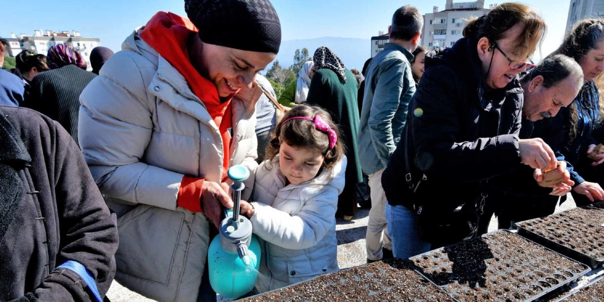 Açık havada, toprak dolu fide viyollerinin başında bir araya gelmiş kadınlar ve çocuklar, bitki yetiştirme etkinliği gerçekleştiriyor. Ön planda, başında siyah bir bere olan gülümseyen bir kadın, yanındaki küçük kız çocuğuyla birlikte mavi bir fısfıs yardımıyla toprağı suluyor. Yanlarında ise gözlüklü bir kadın ve bir erkek, viyollere tohum ekme işlemi yapıyor. Arka planda ise diğer katılımcılar ve şehir binaları görülüyor.