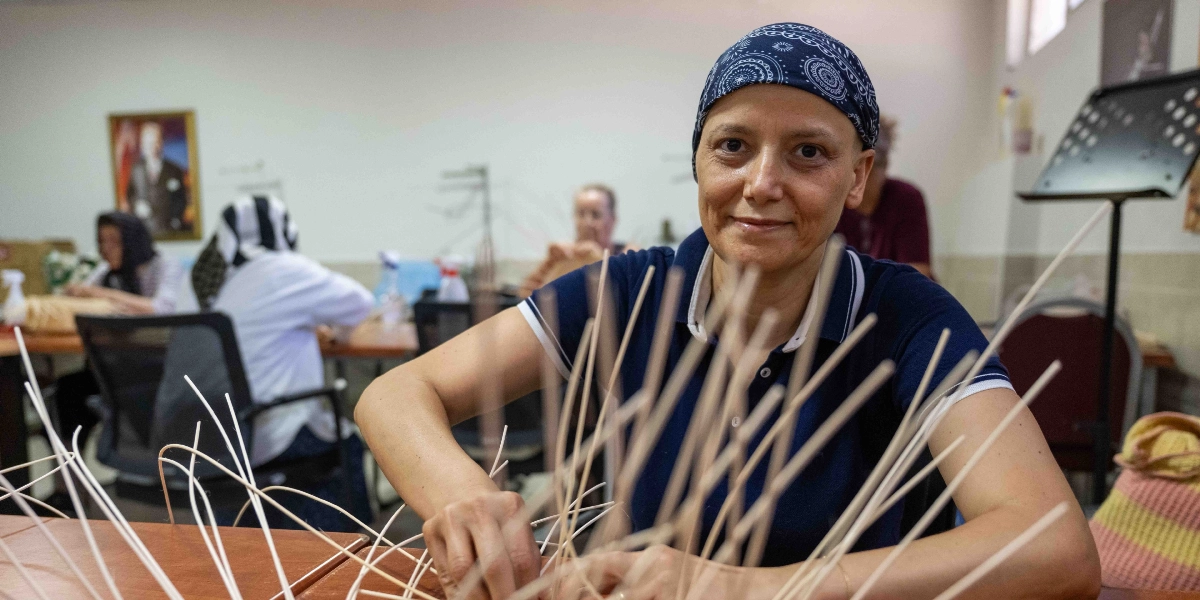 Kafasında lacivert desenli bir bandana takılı olan Yeliz, bir atölye ortamında masanın başında oturmuş, elleriyle ince çubuklardan sepet örme çalışması yaparken kameraya gülümseyerek bakıyor. Arka planda el sanatları kursuna katılan diğer kursiyerler de masalarda çalışmaya devam ediyor.