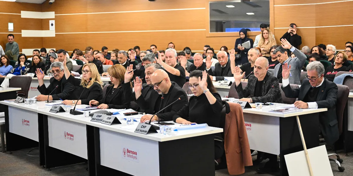 Bornova Belediyesi meclis salonunda düzenlenen toplantıda, masada oturan bir grup katılımcı ellerini kaldırarak oylamaya katılıyor. Ön sırada, üzerinde 