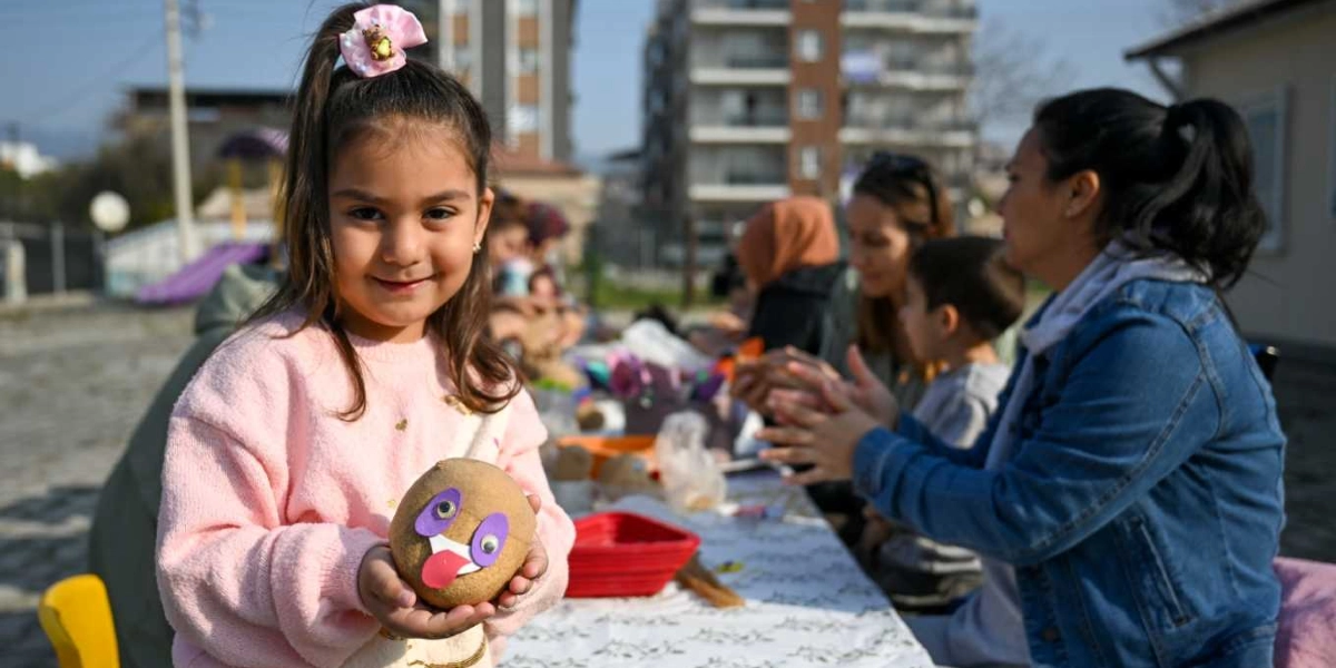 Pembe kazaklı ve saçları tepeden toplanmış küçük bir kız çocuğu, ellerinde tuttuğu kendi yaptığı çim adamı kameraya doğru göstererek gülümsüyor. Arka planda açık havada kurulan bir masada, diğer çocuklar ve yetişkinler atölye çalışmalarına devam ediyor.