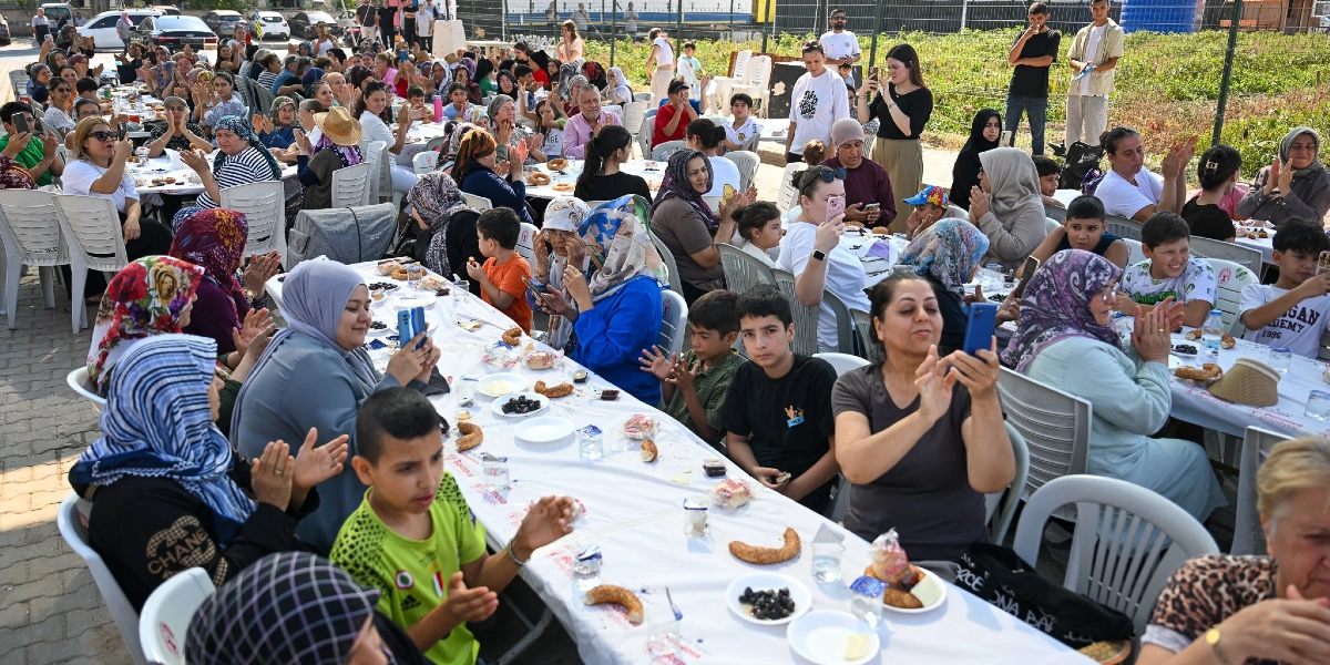 Açık havada, beyaz örtülü uzun masalarda bir araya gelmiş kalabalık bir grup insan, neşeyle alkışlıyor ve cep telefonlarıyla fotoğraf çekiyor. Masaların üzerinde simit, zeytin ve peynir gibi kahvaltılıklar bulunuyor. Çoğunluğu kadın ve çocuklardan oluşan katılımcılar, Bornova Belediyesi logolu örtülerin serili olduğu masalarda otururken, arka planda tel çitlerle çevrili bir tarım alanı ve ağaçlar görülüyor.