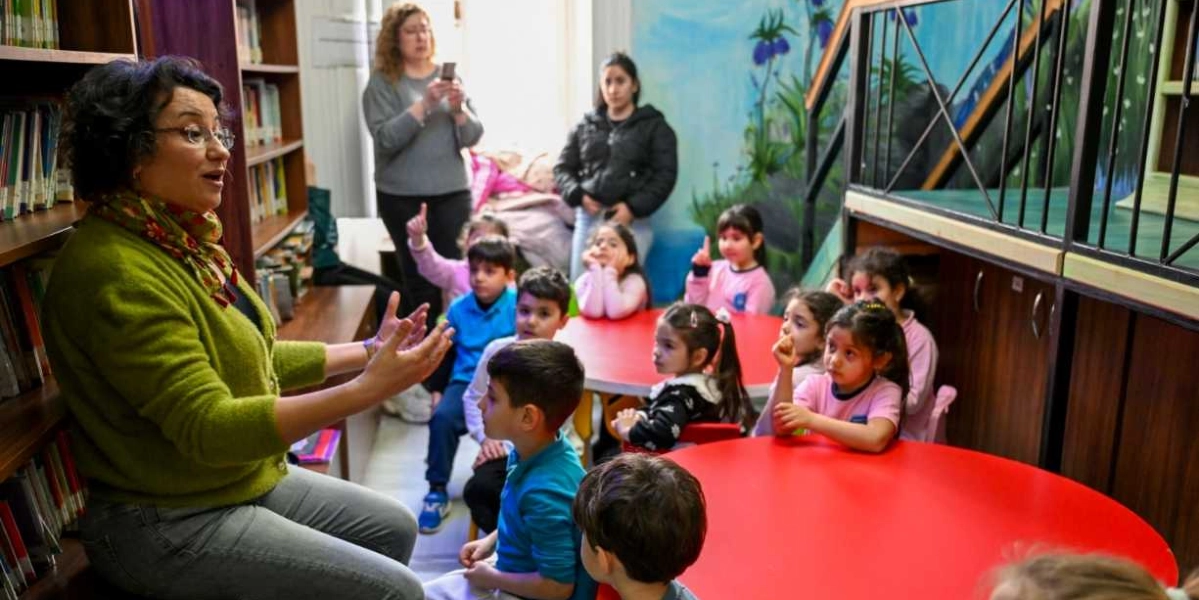 Kütüphanede kitap raflarının önünde oturan ve ellerini kullanarak hikaye anlatan gözlüklü bir kadın, etrafındaki kırmızı masalarda oturan bir grup çocuğa hitap ediyor. Çocuklar ilgiyle kadını dinlerken, arka planda iki yetişkin onları izliyor ve duvarlarda renkli doğa temalı çizimler yer alıyor.