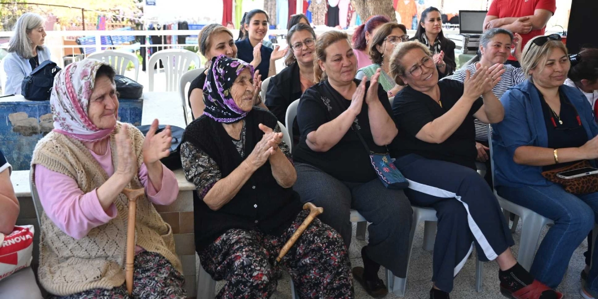 Açık havada düzenlenen bir etkinlikte, farklı yaş gruplarından kadınlar yan yana oturmuş alkışlıyor. Ön sırada, başörtülü ve bastonlu iki yaşlı kadın ile yanlarında oturan orta yaşlı kadınlar neşeyle gülümseyerek ellerini çırpıyor. Arka planda ise etkinliği izleyen diğer katılımcılar ve stantlar yer alıyor.
