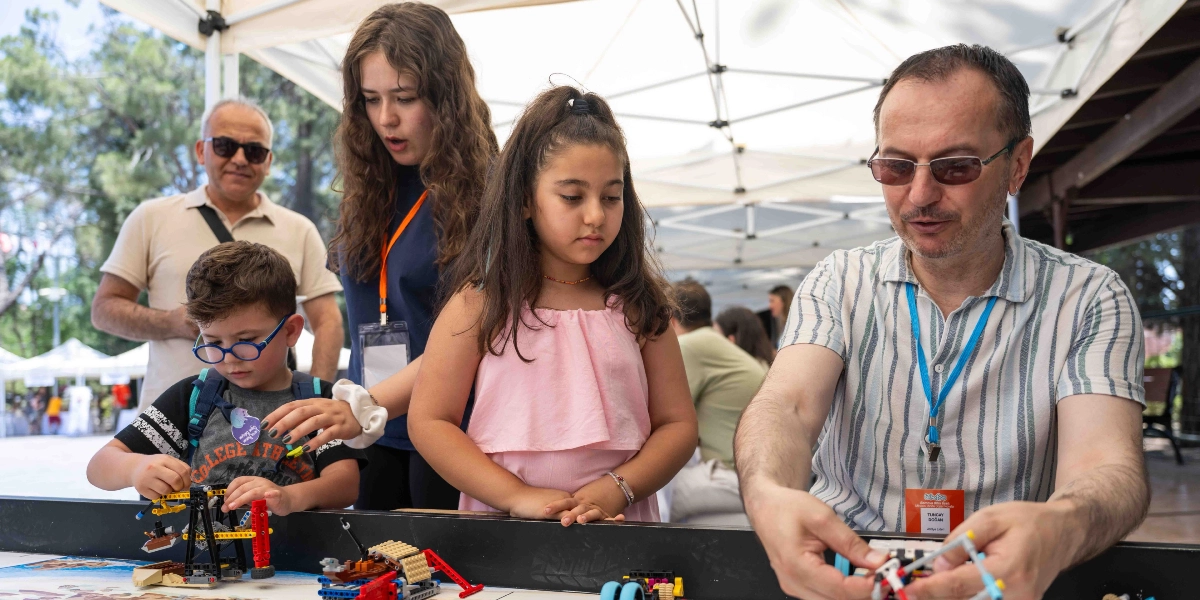 Açık havada, beyaz çadırların altında kurulan bir stantta, yetişkinler ve çocuklar birlikte lego parçalarıyla robotik projeler üzerinde çalışıyor. Ön planda, gözlüklü bir çocuk ve yanında pembe askılı bluz giyen bir kız çocuğu, masanın üzerindeki lego düzeneklerini inceliyor. Sağ tarafta, çizgili gömlekli ve gözlüklü bir yetişkin, elindeki parçalarla bir model oluştururken onlara rehberlik ediyor. Arka planda ise başka bir yetişkin onları izliyor. Katılımcıların boyunlarında etkinlik kartları asılı duruyor.