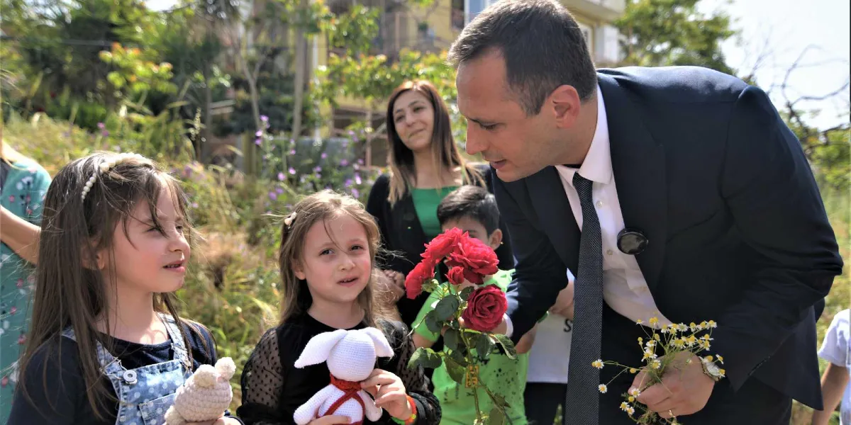 Takım elbiseli bir adam, açık havada bir grup çocuğa kırmızı güller ve papatyalar uzatıyor. Çocuklardan biri elinde örgü bir tavşan oyuncağı tutuyor, arkada ise bir kadın gülümseyerek onları izliyor.