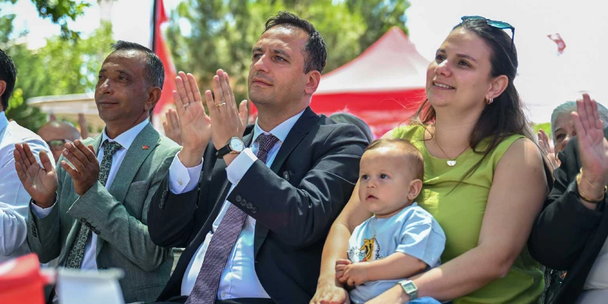 Açık havada düzenlenen bir etkinlikte, takım elbiseli bir erkek ve yanında yeşil bluzlu bir kadın, kucaklarında küçük bir bebekle birlikte alkışlayarak sahneye bakıyor. Yanlarında gri ceketli başka bir erkek daha bulunuyor ve arka planda kırmızı çadırlar ile ağaçlar görülüyor.