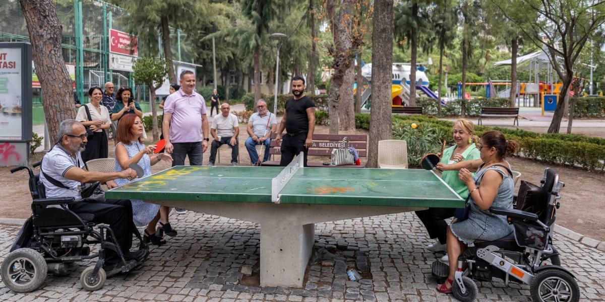 Bornova'da bir parkta, tekerlekli sandalyedeki iki kişi ve yanlarındaki arkadaşlarıyla birlikte masa tenisi oynadıkları keyifli bir an. Yeşil bir masa tenisi masasının iki tarafında oturan oyuncular ellerinde raketlerle topa vurmaya çalışırken, arkalarında onları izleyen ve destekleyen bir grup insan bulunuyor. Arka planda ağaçlar, banklar ve çocuk oyun alanı yer alıyor.