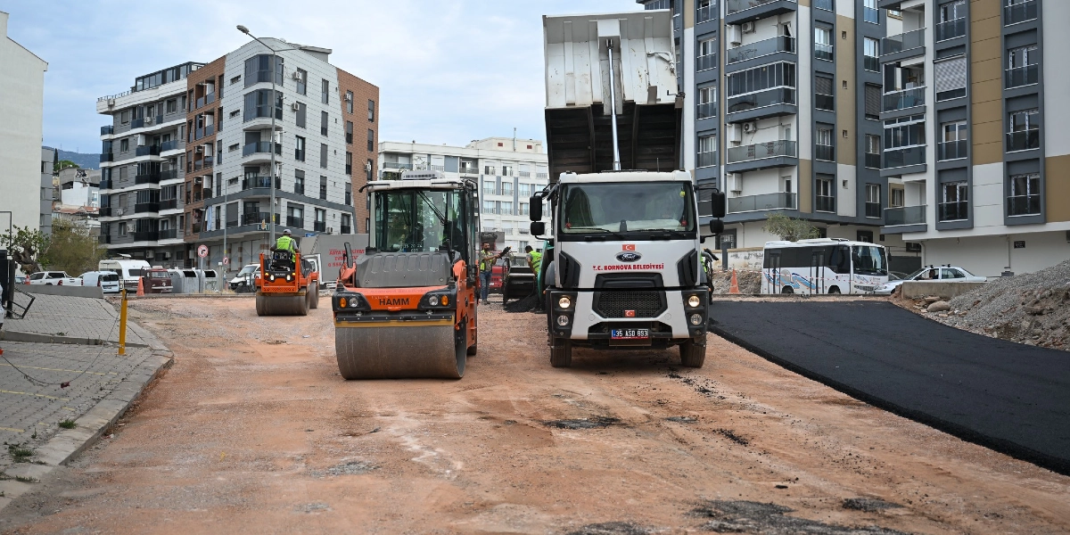 Şehir merkezinde, çok katlı apartmanların arasında devam eden yol yapım çalışmasında, T.C. Bornova Belediyesi'ne ait damperli bir kamyon ve Hamm marka bir silindir iş makinesi toprak zeminde asfaltlama çalışması yapıyor.