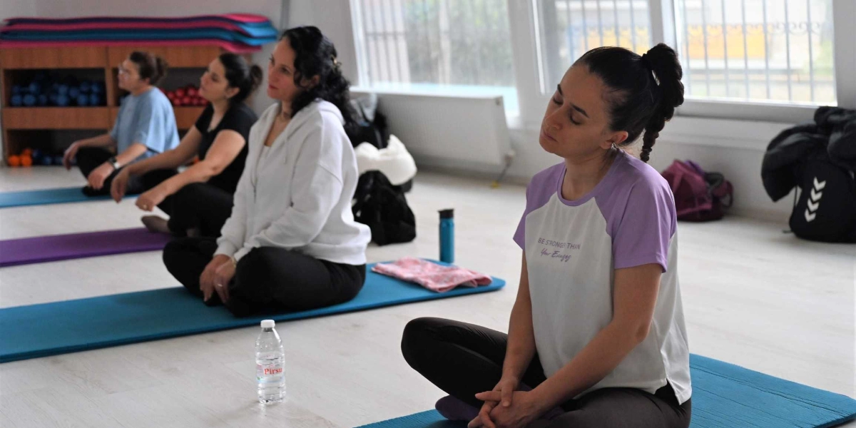 Aydınlık bir spor salonunda, yoga matları üzerinde bağdaş kurarak oturan ve esneme hareketleri yapan dört kadın. Ön plandaki kadın, üzerinde 