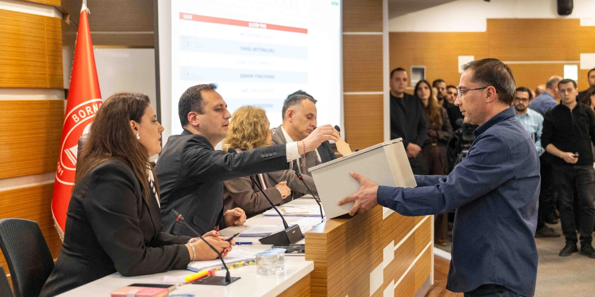 Bornova Belediyesi meclis toplantısında, bir meclis üyesi elindeki oy pusulasını şeffaf oy sandığına atıyor. Masada oturan görevliler süreci takip ederken, arka planda meclis üyeleri ve izleyiciler yer alıyor. Salonun sol tarafında Bornova Belediyesi'nin kırmızı renkli bayrağı, arkadaki projeksiyon ekranında ise isim listeleri görünüyor.