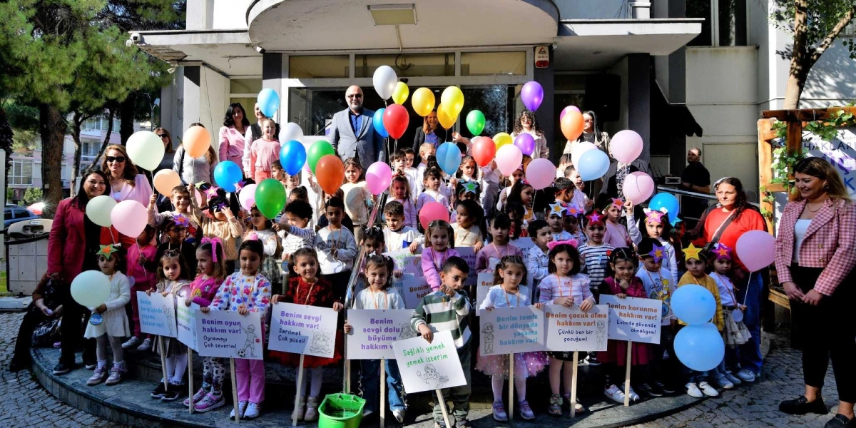 Bornova’da bir bina önünde toplanmış, ellerinde renkli balonlar ve üzerinde çocuk haklarıyla ilgili mesajlar yazılı dövizler tutan çocuklar ve onlara eşlik eden yetişkinler. Çocuklar, 
