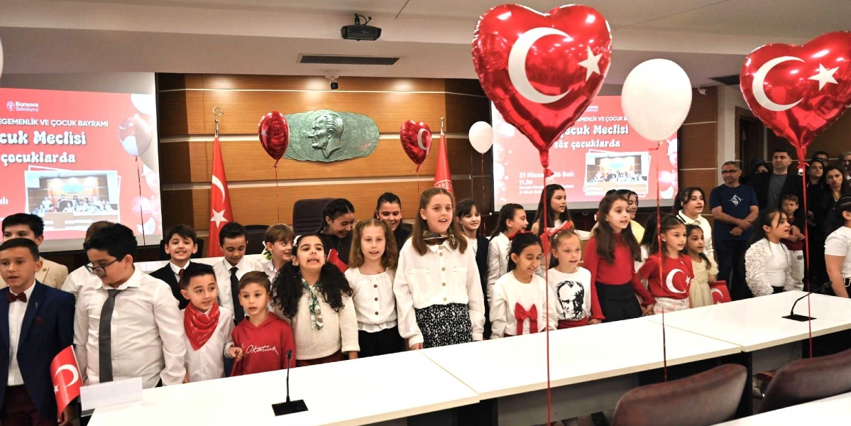 Bornova Belediyesi meclis salonunda, üzerinde Atatürk kabartması bulunan ahşap duvarın önünde duran bir grup çocuk, 23 Nisan Ulusal Egemenlik ve Çocuk Bayramı kutlaması kapsamında şarkı söylüyor. Çocukların bir kısmı beyaz gömlekler, bir kısmı ise kırmızı kıyafetler giymiş durumda; bazıları ellerinde Türk bayrakları tutuyor. Salonun tavanından sarkan kalp şeklinde Türk bayraklı balonlar ve beyaz balonlar ortamı süslüyor. Arka planda yer alan ekranlarda 