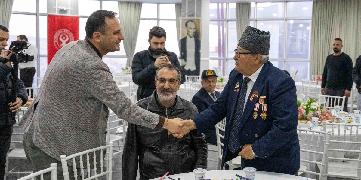 Bornova Belediye Başkanı Mustafa İduğ, üzerinde madalyalar bulunan kıdemli bir gazinin elini sıkarak selamlaşıyor. Arka planda beyaz örtülü masaların bulunduğu bir etkinlik salonunda, bir kameraman çekim yaparken diğer katılımcılar onları izliyor. Duvarlarda Bornova Belediyesi'nin logosunun bulunduğu kırmızı bir bayrak ve Mustafa Kemal Atatürk'ün portresi yer alıyor.