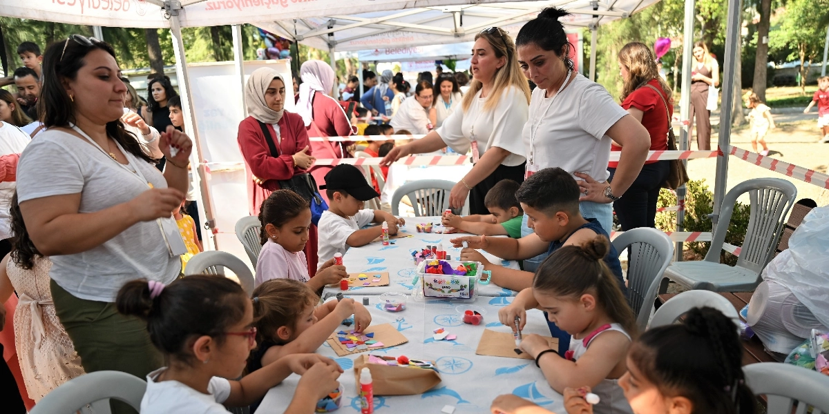 Açık havada, beyaz bir çadırın altında çocuklar masalara oturmuş el işi etkinlikleri yapıyor. Çocuklar, masanın üzerindeki renkli kağıt parçaları, yapıştırıcılar ve çeşitli el işi malzemeleriyle yaratıcı çalışmalar gerçekleştiriyor. Etkinliğe eşlik eden yetişkinler çocuklara yardımcı oluyor. Arka planda parkın yeşil dokusu ve başka katılımcılar görülüyor.