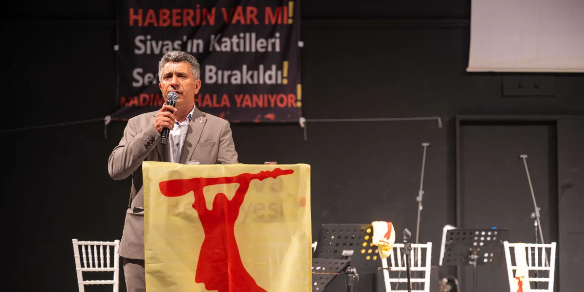 Gri takım elbiseli bir konuşmacı, üzerinde kırmızı bir figürün yer aldığı sarı bir kürsü önünde mikrofonla hitap ediyor. Konuşmacının arkasındaki siyah panoda 