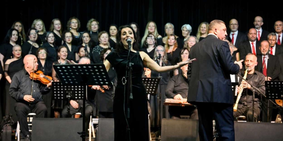 Siyah kıyafetler giymiş kalabalık bir koro, sahnede performans sergiliyor. Ön planda, siyah kadife elbiseli kadın solist mikrofon başında şarkı söylerken, sağ tarafında orkestra şefi sırtı dönük şekilde koroyu yönetiyor. Sol tarafta keman çalan bir müzisyen, sağ tarafta ise klarnet çalan başka bir müzisyen yer alıyor. Arka planda ise çok sayıda korist, siyah bir fon önünde ayakta durarak eşlik ediyor.