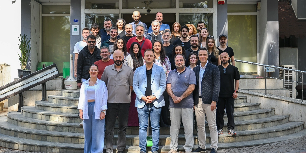 Bornova Belediyesi Şehir Tiyatrosu ekibi, yeni sezon başlangıcı için bir tiyatro binasının girişindeki merdivenlerde toplu halde poz veriyor. Farklı yaş ve tarzlarda giyinmiş kadın ve erkeklerden oluşan kalabalık grup, binanın önünde birkaç sıra halinde dizilmiş şekilde kameraya gülümsüyor.