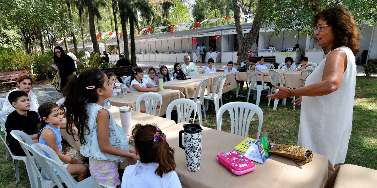 Açık havada, ağaçların altındaki masalarda oturan bir grup çocuğa, ayakta duran gözlüklü bir kadın konuşma yapıyor. Çocuklar masaların etrafında dikkatle dinlerken, masaların üzerinde kitaplar, kalemlikler ve su mataraları bulunuyor. Arka planda ise Bornova Belediyesi'nin düzenlediği kitap günleri kapsamında kurulan stantlar ve Türk bayraklarıyla süslü bir alan yer alıyor.