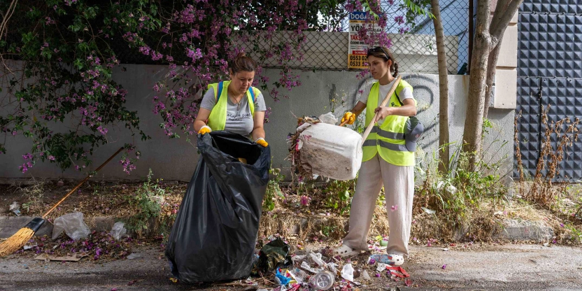 Üzerlerinde neon sarı renkli yelekler bulunan iki kadın, Bornova'da bir sokak kenarında temizlik çalışması yapıyor. Kadınlardan biri elindeki büyük beyaz bir kovayı siyah çöp poşetine boşaltırken, diğeri poşeti açık tutarak ona yardımcı oluyor. Yanlarında uzun saplı bir süpürge ve yerde toplanmayı bekleyen bir miktar çöp bulunuyor. Arka planda mor çiçekli bir ağaç ve üzerinde ilanlar bulunan gri bir duvar yer alıyor.