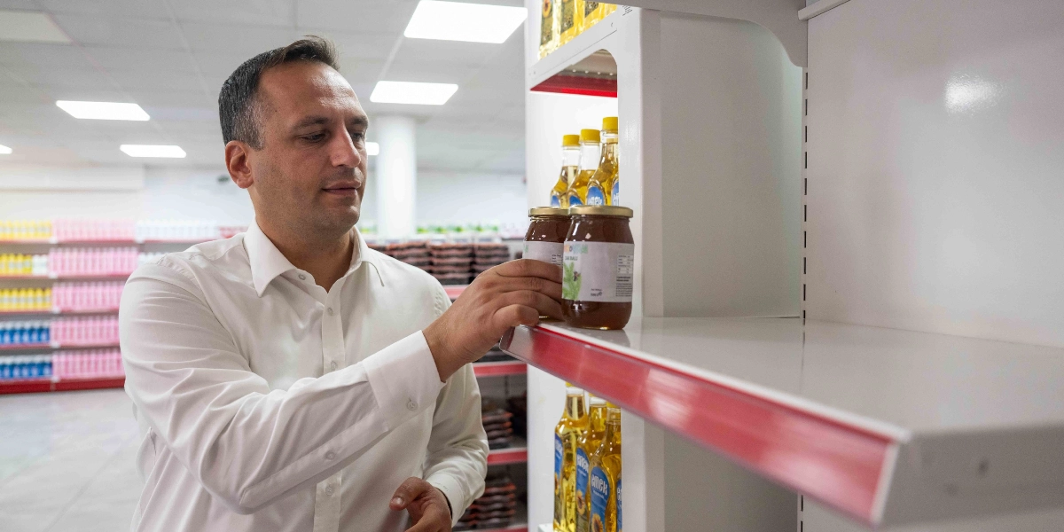 Beyaz gömlekli bir erkek, bir market reyonunda elini rafa uzatarak kavanozda satılan bal ürünlerini inceliyor.