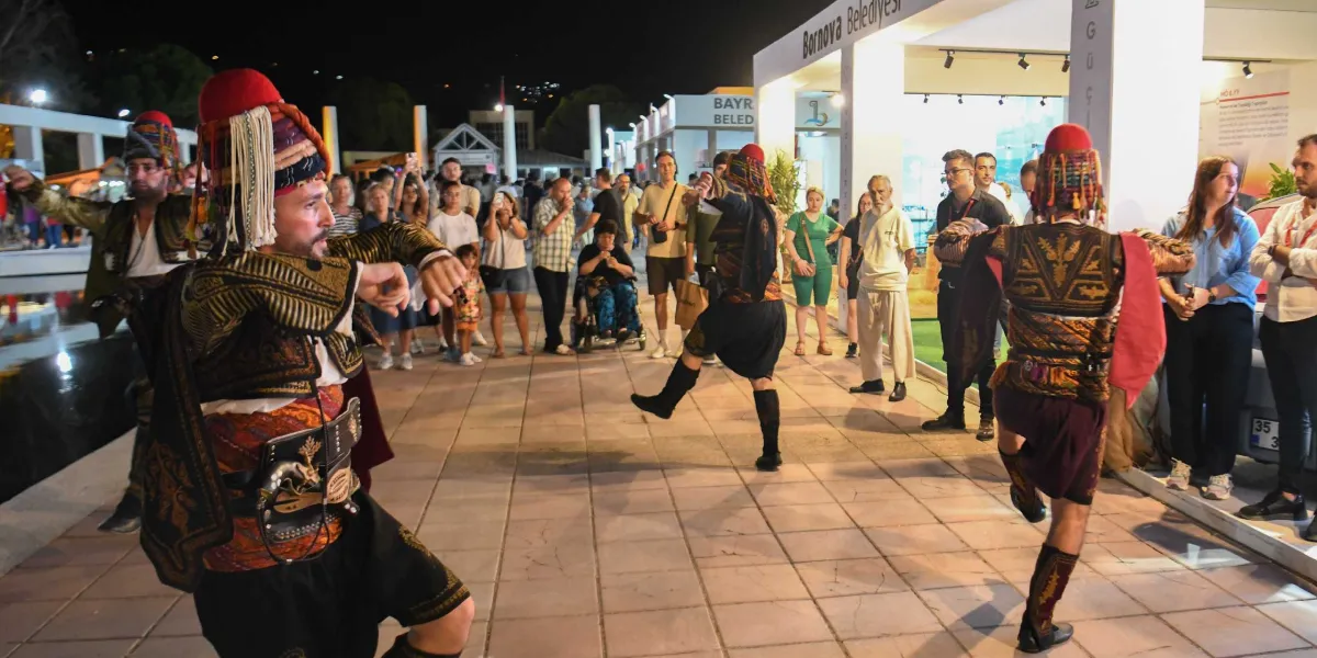 Gece vakti düzenlenen Uluslararası İzmir Fuarı'nda, geleneksel zeybek kıyafetleri giymiş üç erkek dansçı, Bornova Belediyesi standının önünde halk oyunları sergiliyor. Dansçıların etrafında toplanan izleyiciler gösteriyi ilgiyle takip ediyor.