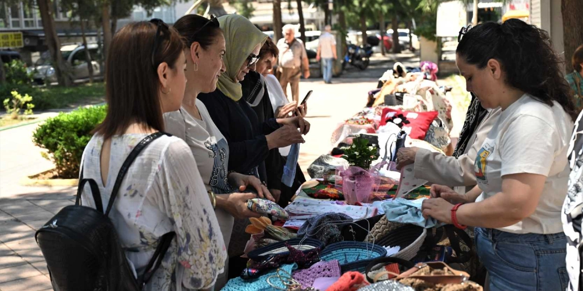 Açık havada kurulan bir stantta el yapımı ürünleri inceleyen kadınlar ve onlara yardımcı olan bir satıcı. Stant üzerinde örgü sepetler, kumaş aksesuarlar ve çeşitli el işi ürünler sergileniyor. Arka planda ağaçlıklı bir yürüyüş yolu ve yürüyen insanlar görünüyor.