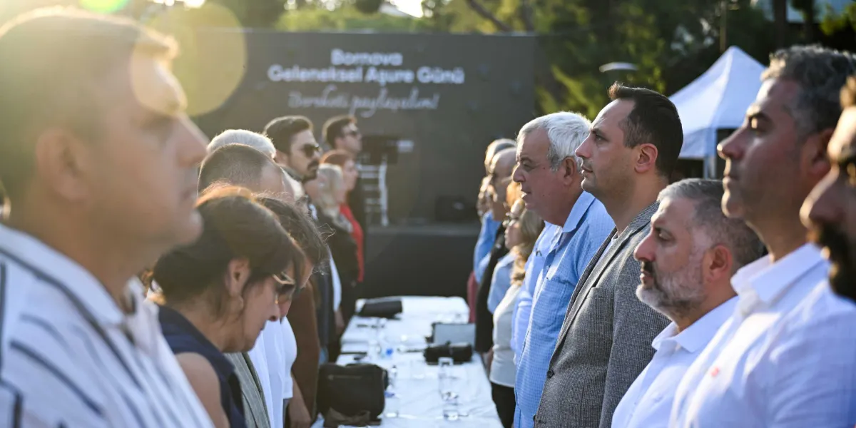 Açık havada düzenlenen Bornova Geleneksel Aşure Günü etkinliğinde, uzun bir masa etrafında yan yana dizilmiş ve saygı duruşunda bulunan bir grup insan. Arka planda üzerinde 
