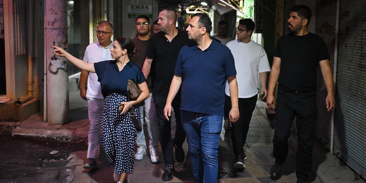 Gece vakti Çamdibi sokaklarında yürüyen bir grup yetkili, altyapı çalışmalarını yerinde inceliyor. Lacivert gömlekli bir kadın eliyle bir noktayı işaret ederek yanındaki gruba bilgi veriyor. Grubun önünde lacivert polo yaka tişört ve kot pantolon giyen bir erkek ile yanında beyaz tişörtlü, gözlüklü bir erkek ve siyah kıyafetli diğer yetkililer yer alıyor. Arka planda kapalı dükkan kepenkleri ve sokak dokusu görülüyor.