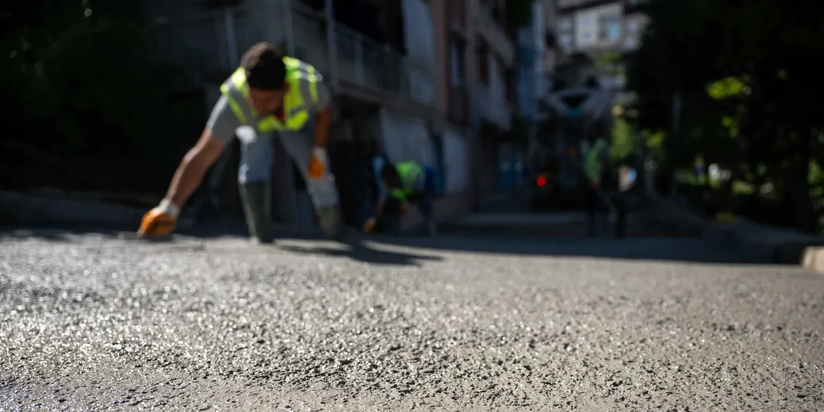 Sarı reflektörlü yelek giyen bir işçi, yokuş bir sokakta yeni dökülen beton zemini mala ile düzeltiyor. Arka planda bulanık şekilde diğer işçiler ve bir beton mikseri görülüyor.