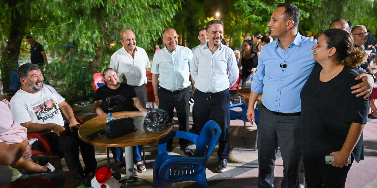 Bornova Belediye Başkanı Ömer Eşki, akşam saatlerinde Çamdibi'nde vatandaşlarla bir araya gelerek sohbet ediyor. Açık havada, ağaçlıklı bir alanda bulunan yuvarlak bir masanın etrafında oturan ve ayakta duran bir grup insan, Başkan Eşki ile samimi bir şekilde iletişim kuruyor. Başkan Eşki, mavi gömlek giymiş bir şekilde yanındaki kadınla gülümseyerek konuşurken, çevresindeki diğer kişiler de keyifli bir atmosferde sohbete eşlik ediyor.