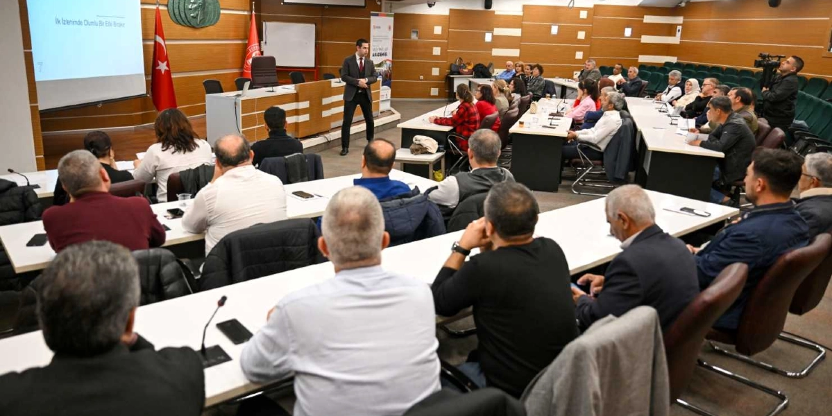 Modern bir konferans salonunda, takım elbiseli bir eğitmen ayakta sunum yaparken, masalarda oturan kalabalık bir katılımcı grubu onu dinliyor. Salonun ön tarafında üzerinde 