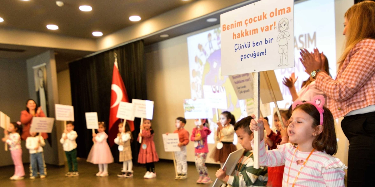 Kapalı bir salonda, ellerinde üzerinde çocuk haklarıyla ilgili sloganlar yazılı dövizler taşıyan bir grup çocuk, Çocuk Hakları Günü'nü kutluyor. Ön planda, üzerinde 