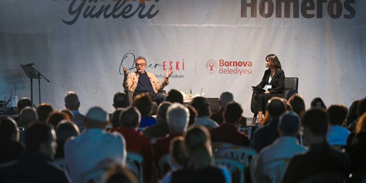 Bornova Belediyesi tarafından düzenlenen bir etkinlikte, sahnede konuşma yapan gözlüklü bir erkek ve onu dinleyen bir moderatör, kalabalık bir izleyici kitlesine hitap ediyor. Arka planda 