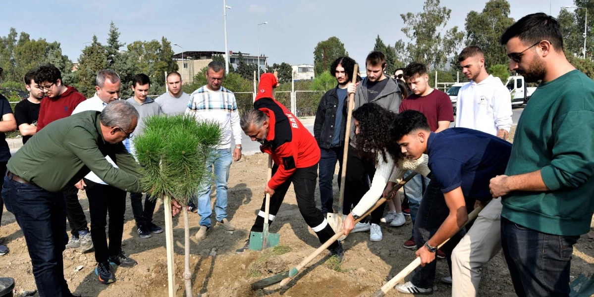 Açık havada, toprak bir alanda bir grup genç ve yetişkinin bir araya gelerek fidan diktiği etkinlik. Kırmızı ceketli bir görevli elindeki kürekle toprağı kazarken, yanındaki gençler ve yetişkinler dikkatle onu izliyor. Ön planda, yeşil yapraklı genç bir çam fidanı dikilmeye hazır bekliyor. Çevredeki katılımcılar günlük kıyafetler içinde, ağaçlandırma çalışmasına destek veriyor.