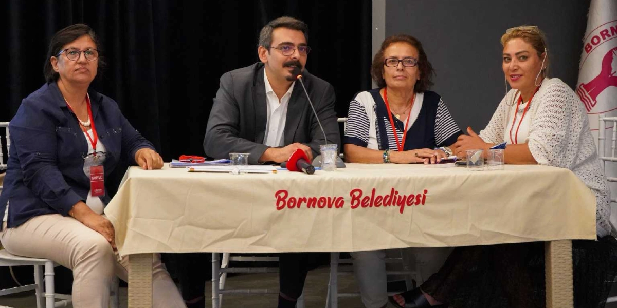 Üzerinde Bornova Belediyesi yazılı bir masanın arkasında yan yana oturan dört kişi, bir toplantı veya panel sırasında yer alıyor. Masanın ortasında mikrofon bulunurken, katılımcıların boyunlarında kırmızı renkli yaka kartı askıları görülüyor. Arka planda ise Bornova Kent Konseyi logosunun yer aldığı bir bayrak bulunuyor.