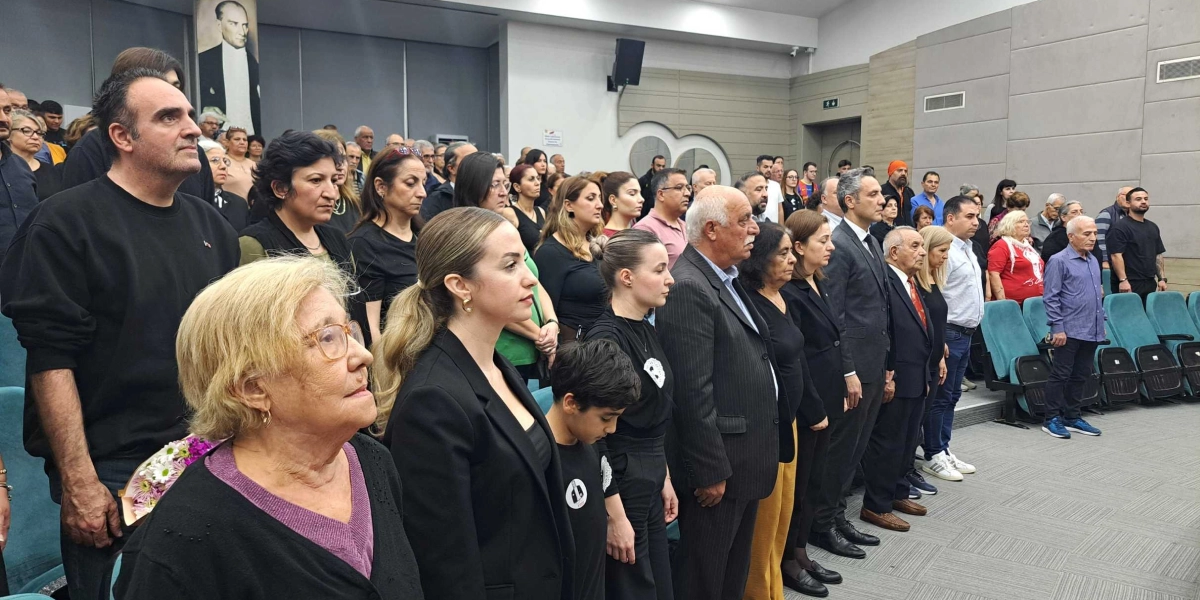 Modern bir konferans salonunda, ayakta duran ve saygı duruşunda bulunan kalabalık bir grup insan. Katılımcıların çoğu siyah kıyafetler giymiş, ciddi ve hüzünlü bir ifadeyle karşıya bakıyor. Salonun ön duvarında, Atatürk'ün büyük bir portresi asılı. Ön planda yaşlı bir kadın, yanında genç bir kadın ve bir çocuk bulunuyor; arka planda ise farklı yaş gruplarından çok sayıda kişi sıralar halinde yer alıyor.